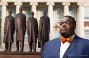 Headshot of Andre Taylor, wearing a blue suit jacket and orange bow tie, standing in front of a blurred image of 4 statues.