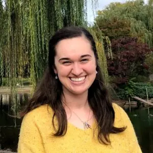 A young woman with long brown hair, wearing a yellow sweater, smiles in front of a pond and lush green trees, including a willow tree, on a bright day.