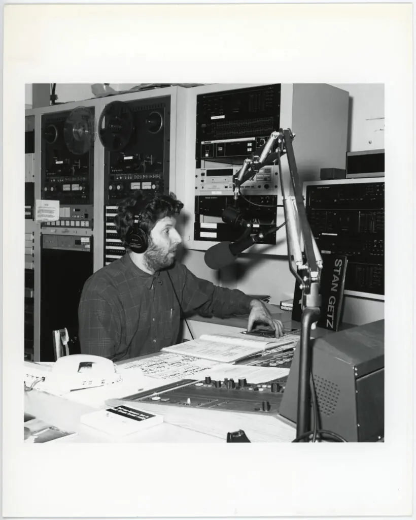 A man wearing headphones speaks into a microphone in a radio station studio, surrounded by audio equipment, control panels, and papers on the desk.