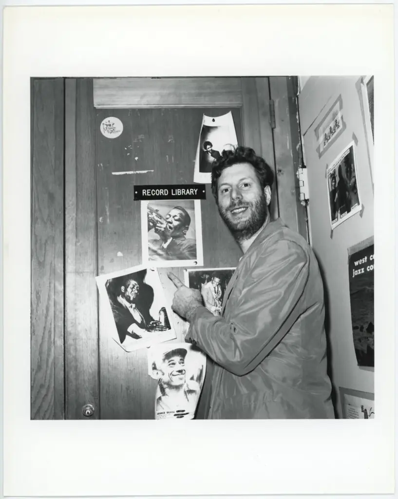 A smiling man in a jacket points at jazz musician photos taped to a wooden door labeled Record Library. More photos and posters are taped to the adjacent wall.
