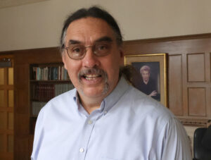 A man with long dark hair, glasses, and a mustache smiles while standing indoors in front of bookshelves and a framed portrait on a wood-paneled wall.