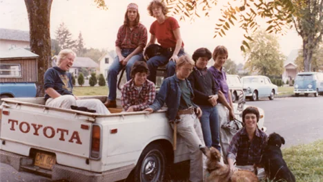 A group of eight people, mostly young adults, and two dogs sit and stand on the back of a white Toyota pickup truck parked on a residential street lined with trees and vintage cars.
