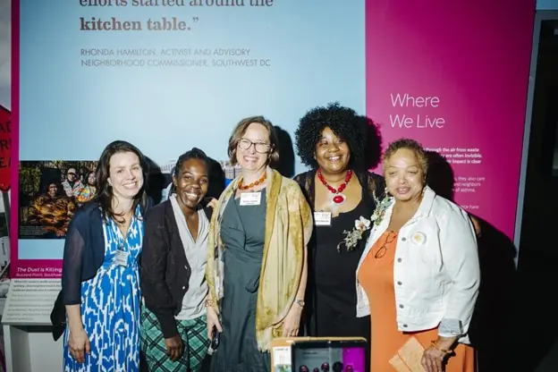 Five women stand together smiling in front of a colorful exhibit display, wearing a mix of professional and festive attire. A quote and exhibit text are visible behind them.