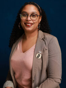 A smiling woman with glasses, curly hair, and hoop earrings wears a tan blazer over a light pink top. She has a “VOTE” button pinned to her blazer and sits in front of a dark blue background.
