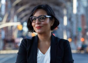 A woman with short dark hair, glasses, and a black blazer stands outside with a confident smile. The background is an urban street scene with blurred lights and structures.