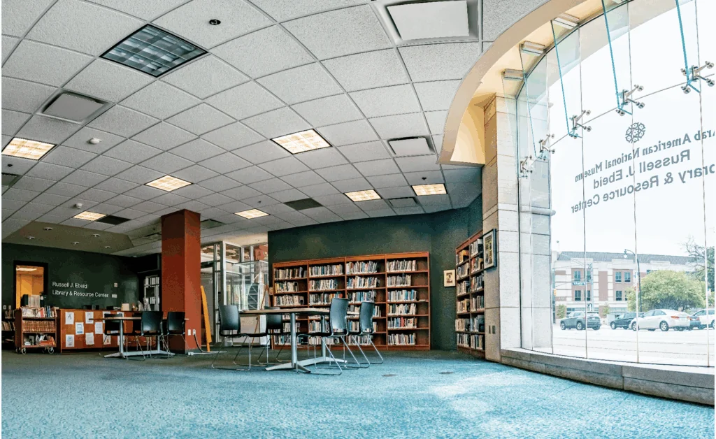 A modern library room with bookshelves, tables, and chairs sits beneath a curved ceiling. Large windows let in natural light and offer a view of buildings outside. The floor is carpeted in blue.
