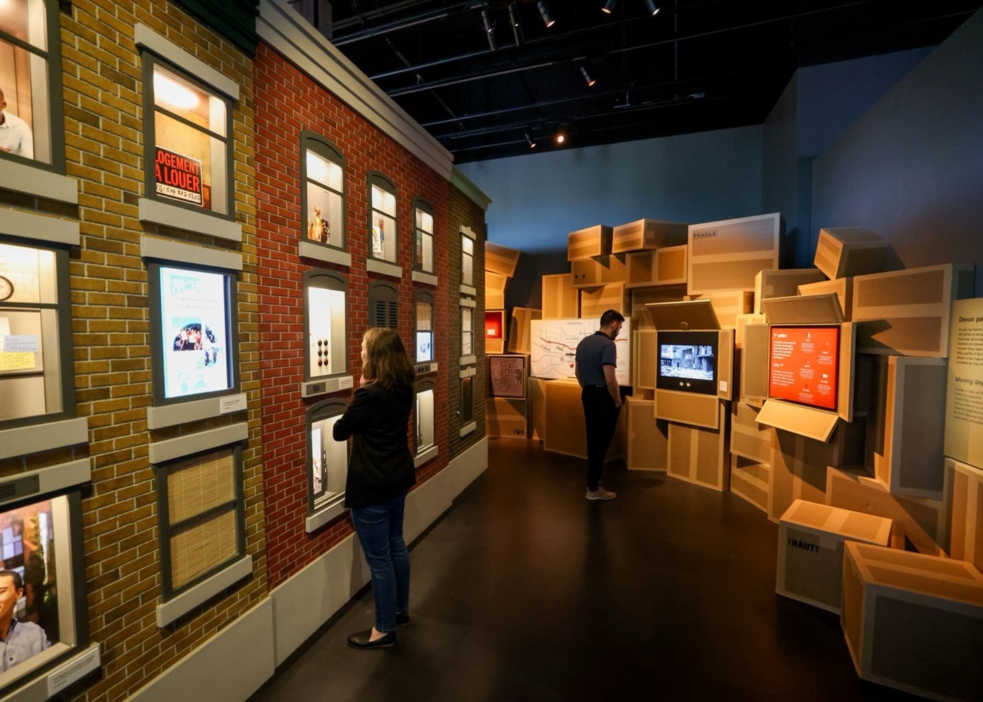 Two people view interactive displays and exhibits on brick and box-styled walls in a modern, dimly lit museum or gallery setting.