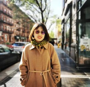 A woman wearing a tan coat, purple sunglasses, and a green patterned scarf stands smiling on a sunny city street with trees, parked cars, and brick buildings in the background.