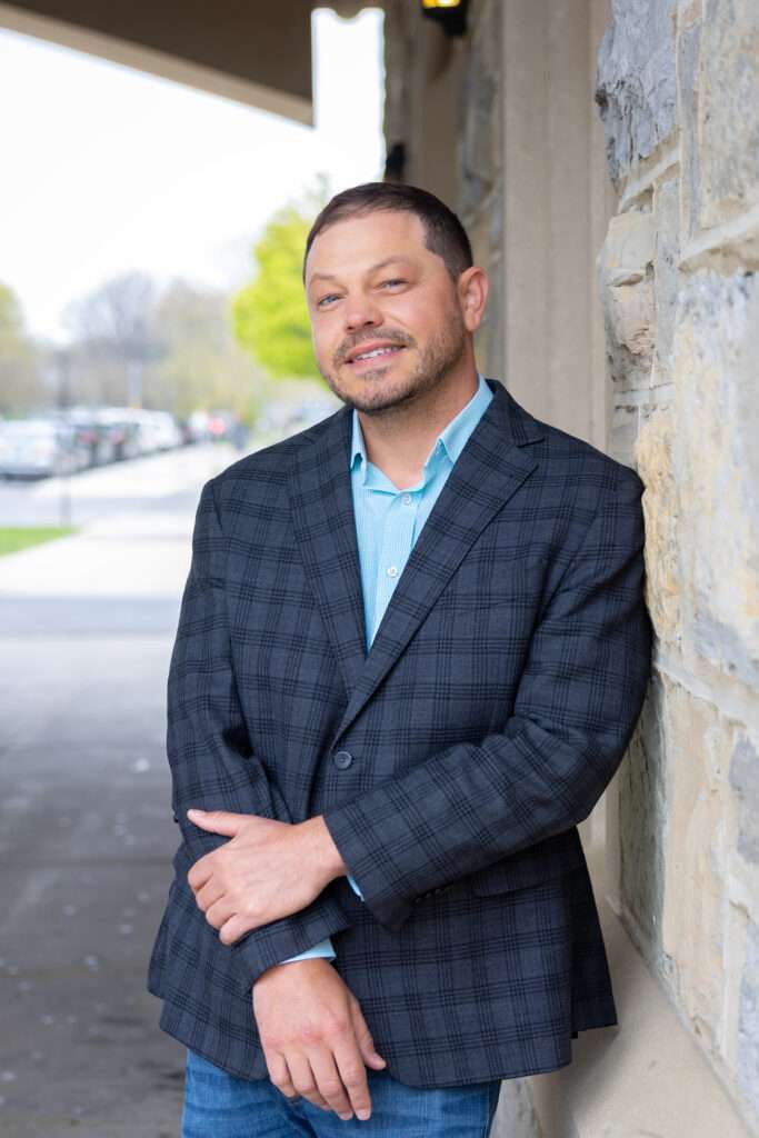 A man in a plaid blazer and blue shirt leans against a stone wall outdoors, smiling with arms crossed. There are trees and parked cars visible in the blurred background.