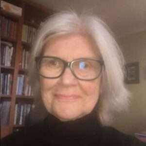 A woman with shoulder-length gray hair and glasses smiles at the camera. She is wearing a black turtleneck and is in a room with bookshelves in the background.