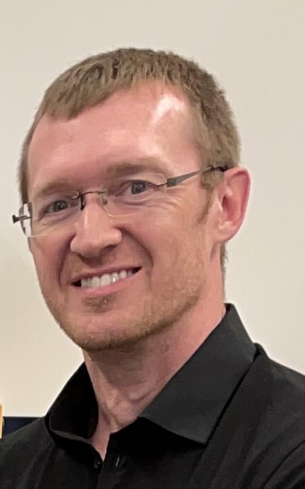 A man with short brown hair, glasses, and a trimmed beard smiles at the camera. He is wearing a black collared shirt and is in front of a plain light-colored background.
