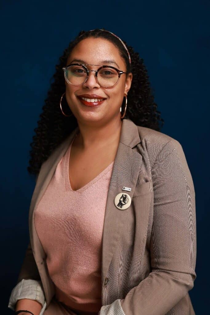 A woman with curly hair and glasses, wearing a beige blazer, pink top, and hoop earrings, smiles in front of a dark blue background. She has a round pin on her lapel.