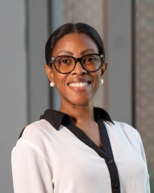 A woman wearing glasses and a white blouse with a black collar smiles at the camera. She has her hair pulled back and is standing indoors with a blurred background.