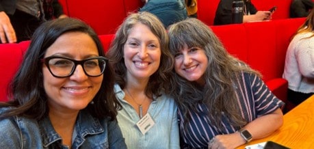 Three women smiling and sitting together at a wooden table with red booth seating in the background. One woman is wearing glasses and another has a name tag.