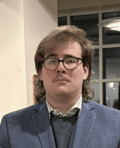 A person with light brown hair, glasses, a mustache, and a blue checked blazer over a sweater and collared shirt stands indoors with windows in the background.