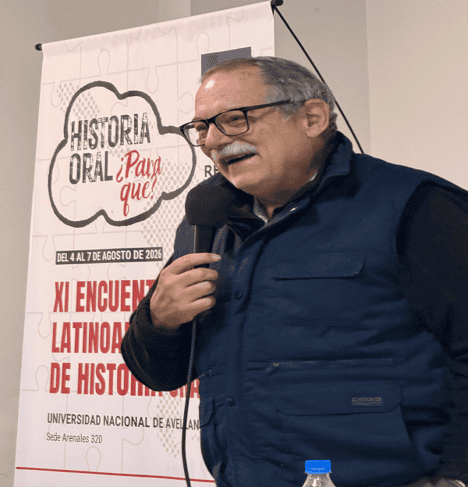An older man in glasses and a blue vest speaks into a microphone at a history event. A poster behind him reads Historia Oral ¡Para Qué! and announces the XI Encuentro Latinoamericano de Historia Oral.