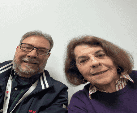 Two adults, a man with glasses and a beard wearing a jacket, and a woman with short brown hair wearing a purple sweater, smiling and posing for a selfie in front of a plain white wall.